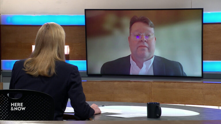 Frederica Freyberg sits at a desk on the Here & Now set and faces a video monitor showing an image of John Jagler.