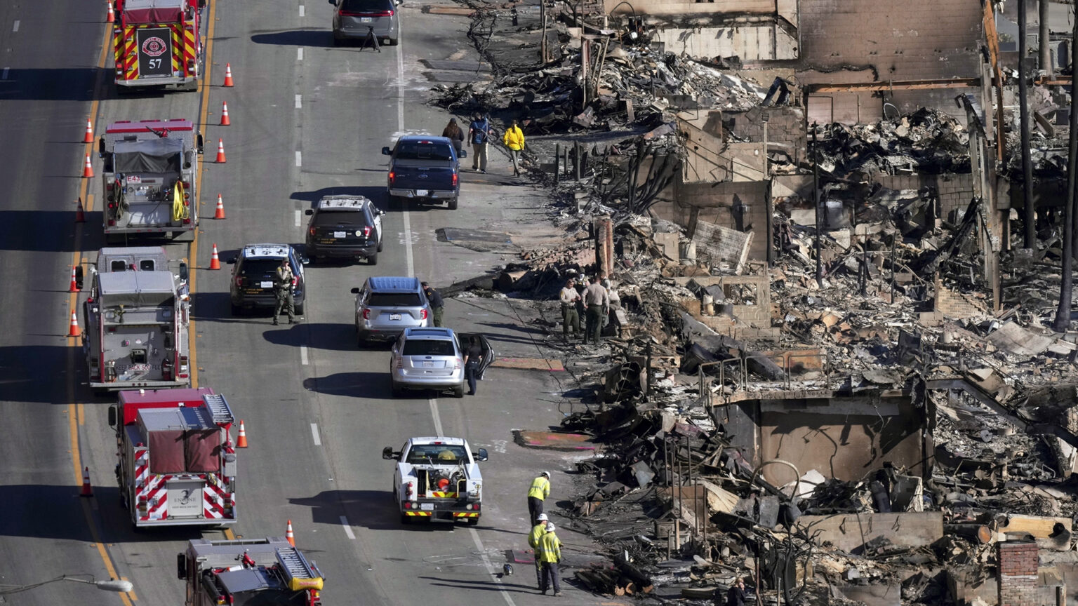 Multiple fire engines are parked in the turn lane of a multi-lane road, with other vehicles parked on the two right-hand lanes next to the ruins of structures destroyed by fire.