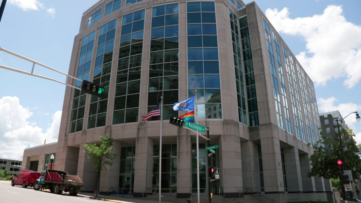 The U.S., Wisconsin and Pride flags fly on flagpoles in front of a multi-story stone and glass office building at an intersection with multiple traffic signal lights and street sights, with other buildings, parked vehicles and trees on either side and in the background.