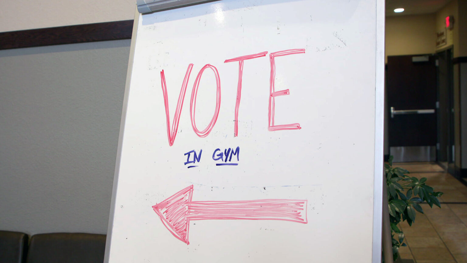 A handwritten sign with the words Vote and In Gym and an arrow are visible on a whiteboard standing in a hallway, with a perpendicular hallway leading toward a commercial metal door with a push bar and illuminated EXIT sign above it.