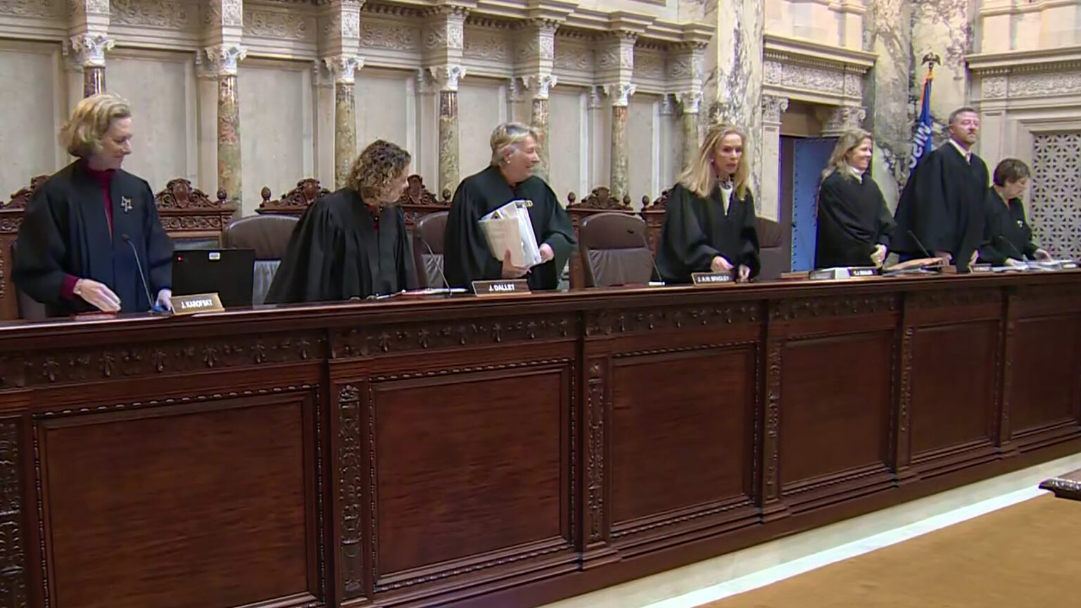 From left to right, Jill Karofsky, Rebecca Dallet, Ann Walsh Bradley, Annette Ziegler, Rebecca Bradley, Brian Hagedorn and Janet Protasiewicz stand at a judicial dais in front of a row of high-backed leather chairs, with another row of high-backed wood and leather chairs behind them, in a room with marble masonry and a Wisconsin flag.