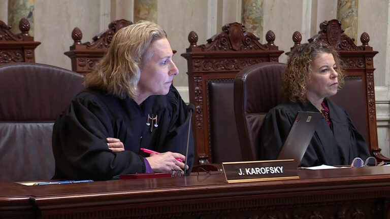 Jill Karofsky and Rebecca Dallet sit in high-backed leather chairs at a judicial bench with a nameplate reading J. Karofsky, with a row of empty high-backed wood and leather chairs behind them in a room with marble pillars and masonry.