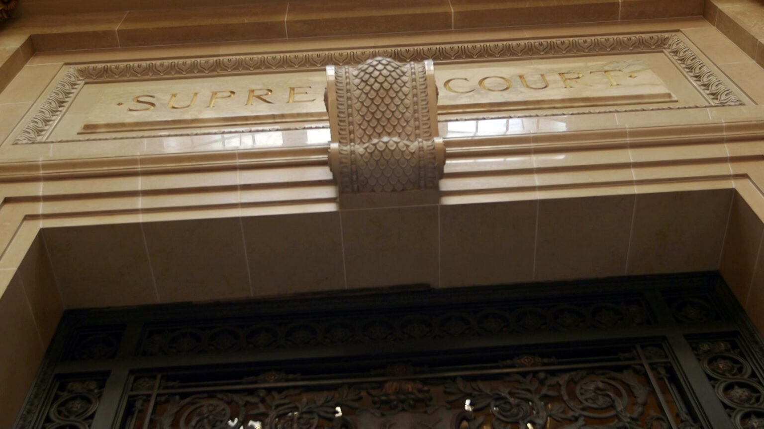 An inlaid carved marble sign reading Supreme Court marks the top of a doorway with a wrought iron filigree and glass door.