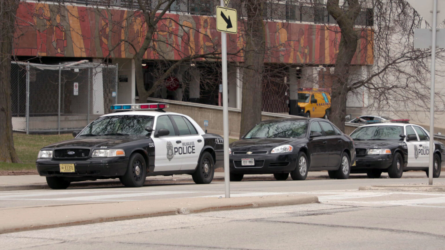 Two Milwaukee Police Department cars and a third vehicle are parked on the side of a street with a concrete median with two street sign poles and a crosswalk, with leafless trees and a multi-story building with a garage entrance in the background.