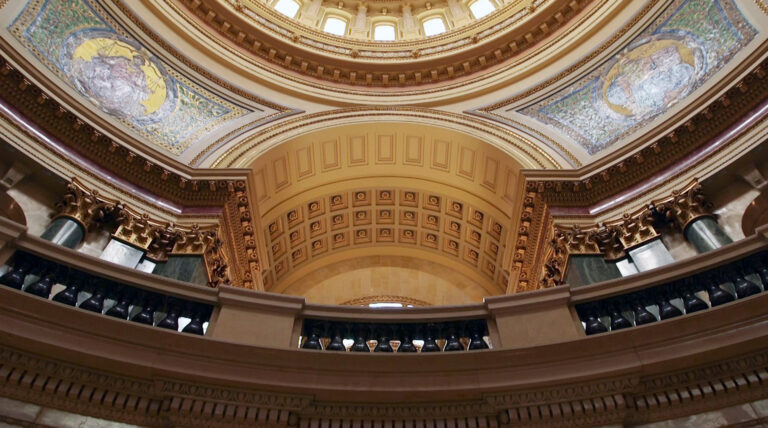 Sunlight reflects off two mosaics depicting Classical figures — one holding scales and labeled Justice and another holding a tablet and labeled Legislation — inside a rotunda beneath a ring of arched windows at the base of a dome, as seen from below inside a building with marble pillars, railings and other masonry in a Beaux-Arts design.