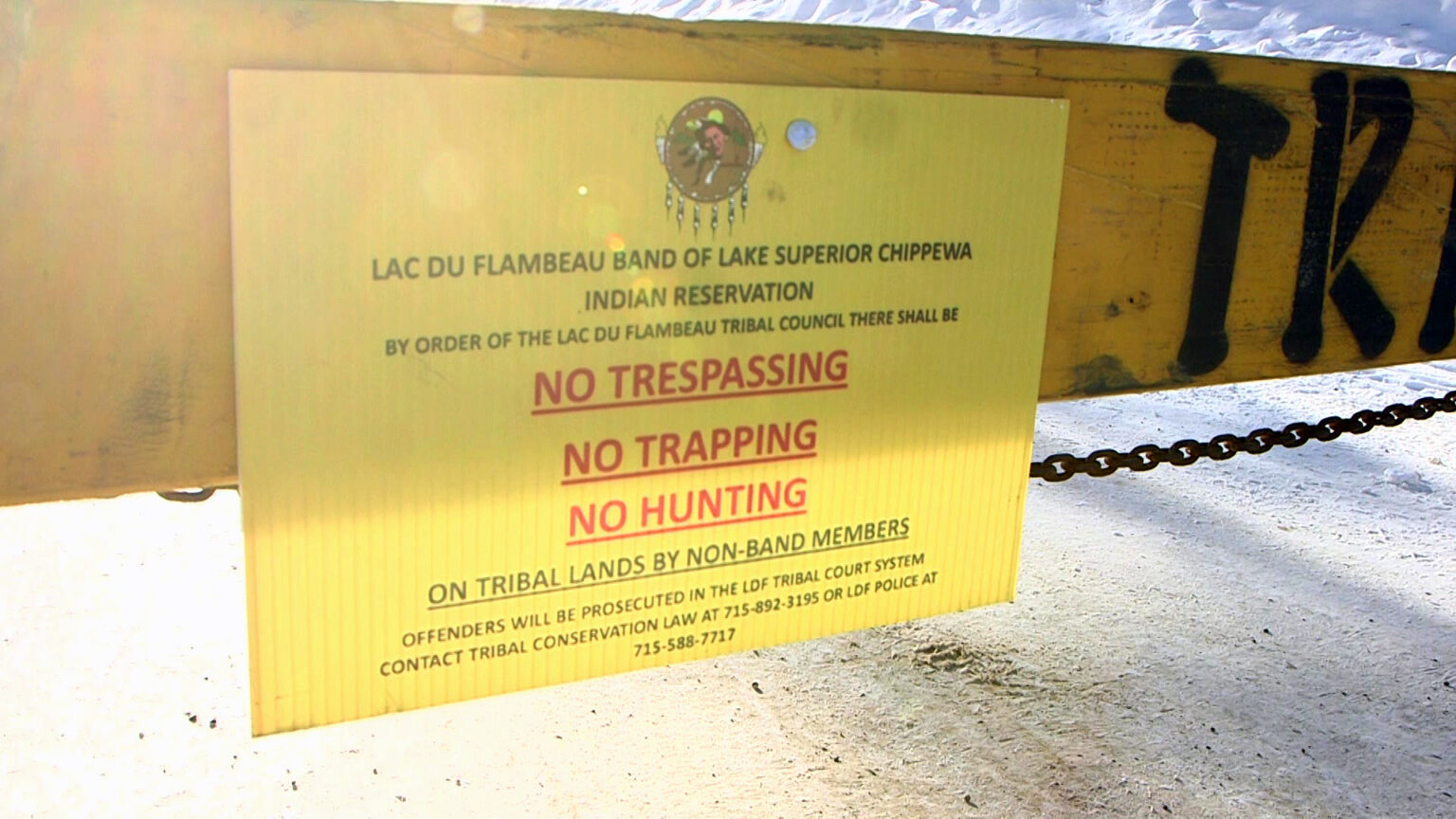 A printed sign with the seal of the Lac du Flambeau Band of Lake Superior Chippewa above the words Lac du Flambeau Band of Lake Superior Chippewa Indian Reservation, By Order of the Lac du Flambeau Tribal Council There Shall Be No Trespassing, No Trapping, No Hunting on Tribal Lands by Non-Band Members, Offenders Will Be Prosecuted in the LDF Tribal Court System and contact information is attached to a wood road barrier with a metal chain hanging underneath it above a road covered in snow.