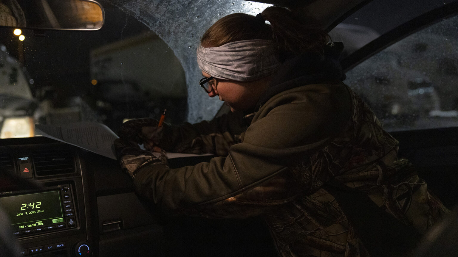 Sandy Hawn writes on a sheet of paper on the dashboard on the passenger front seat of a car with a digital screen below the windshield showing a time of 2:42 on Jan. 9, 2025.