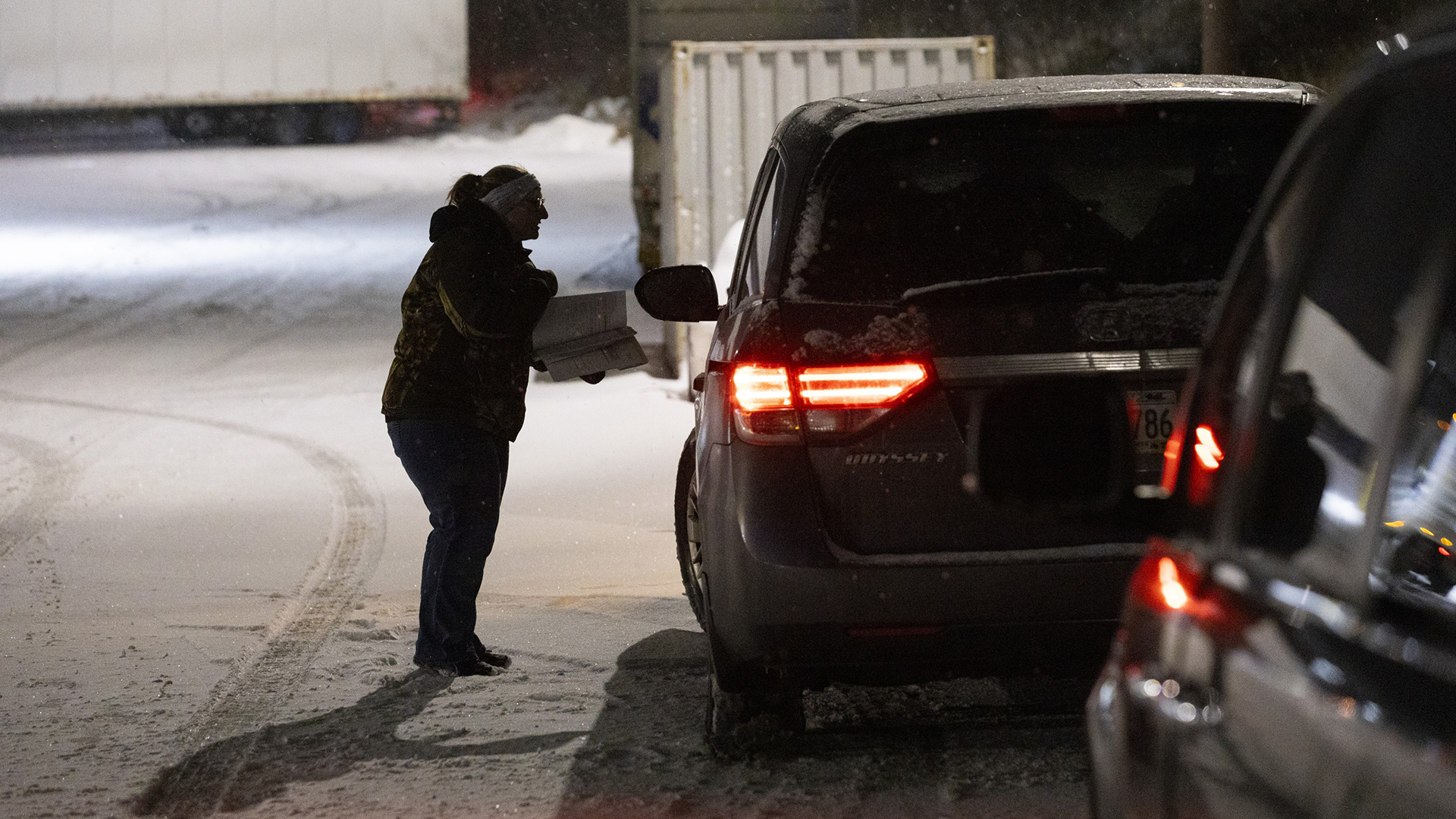 Sandy Hahn stands next to the driver's side window of a parked vehicle with illuminated rear lights in a snow-covered parking lot at night, with metal garbage container and a parked truck trailer in the background.