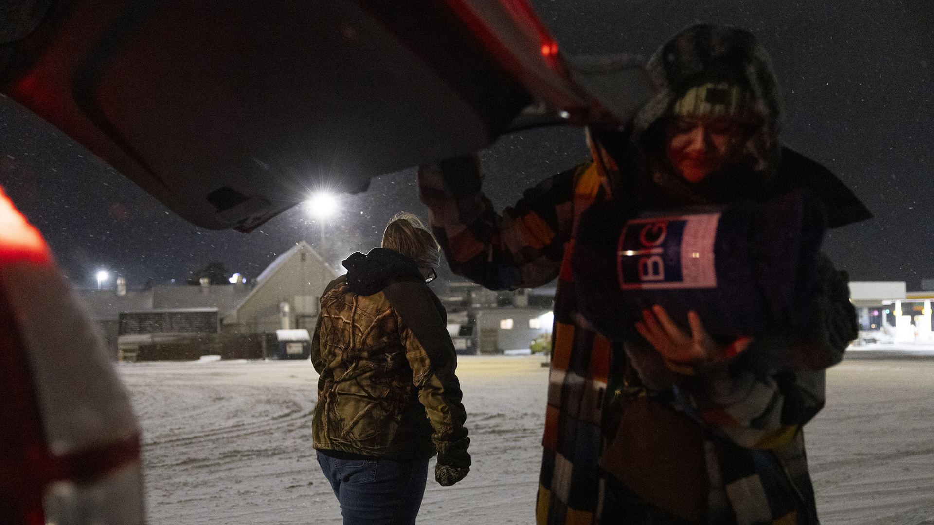 Britanie Peaslee holds a rolled blanket inside its retail packaging in her left hand while using her right hand to lower and close the rear hatch of a vehicle, with Sandy Hahn walking away in the background on a snow-covered parking lot with buildings in the background.