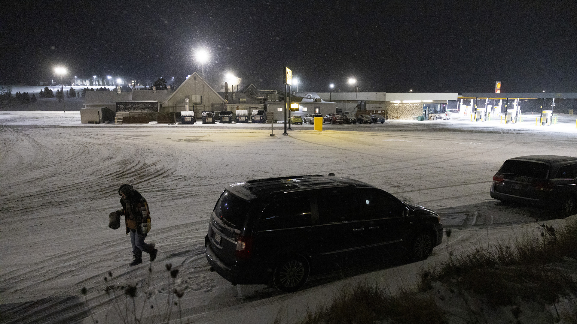 Britanie Peaslee walks away from two vehicles parked at the edge of a snow-covered parking lot covered with tire tracks, with a gas station and adjacent buildings illuminated by light poles in the background as snow falls under a night sky.