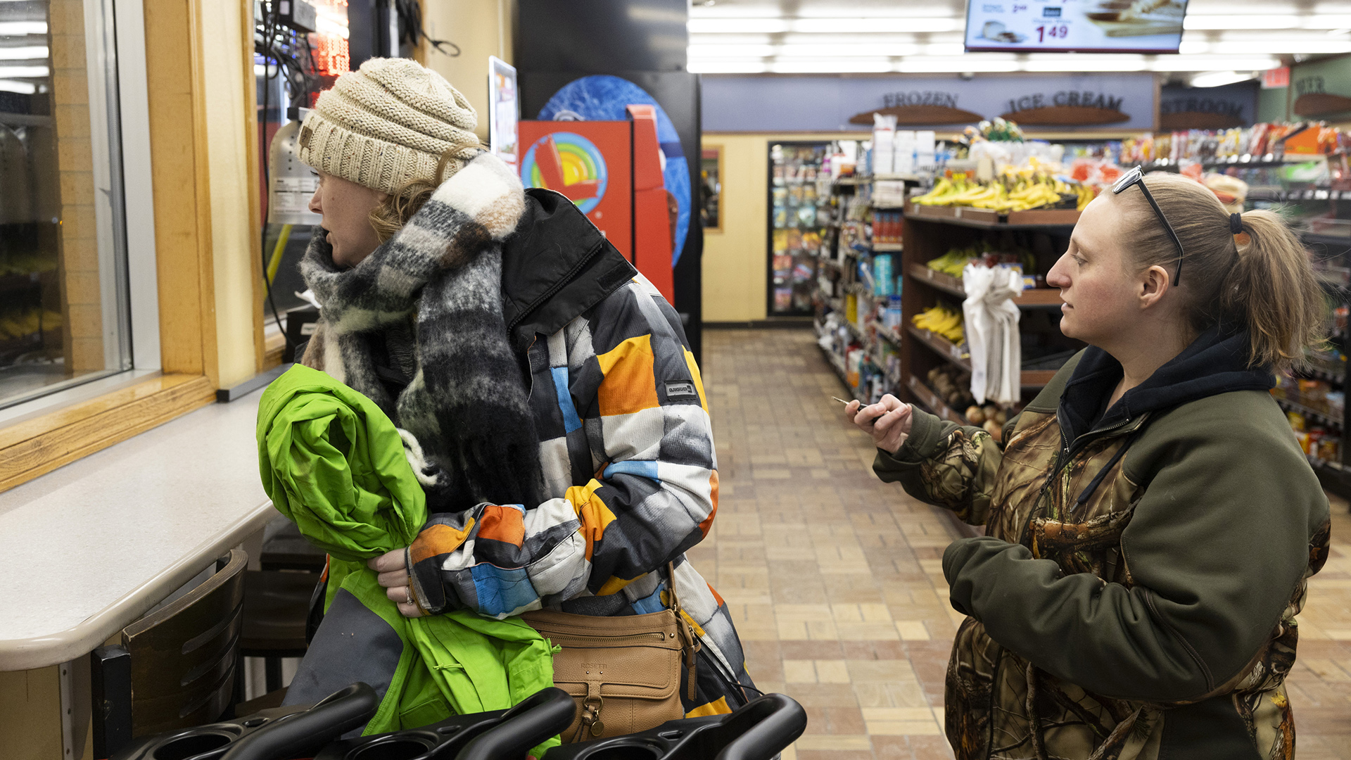 Britanie Peaslee and Sandy Hahn stand next to a row of shopping cars and look out a window in a convenience store with rows of product-covered shelves, a brick floor and overhead fluorescent lighting.