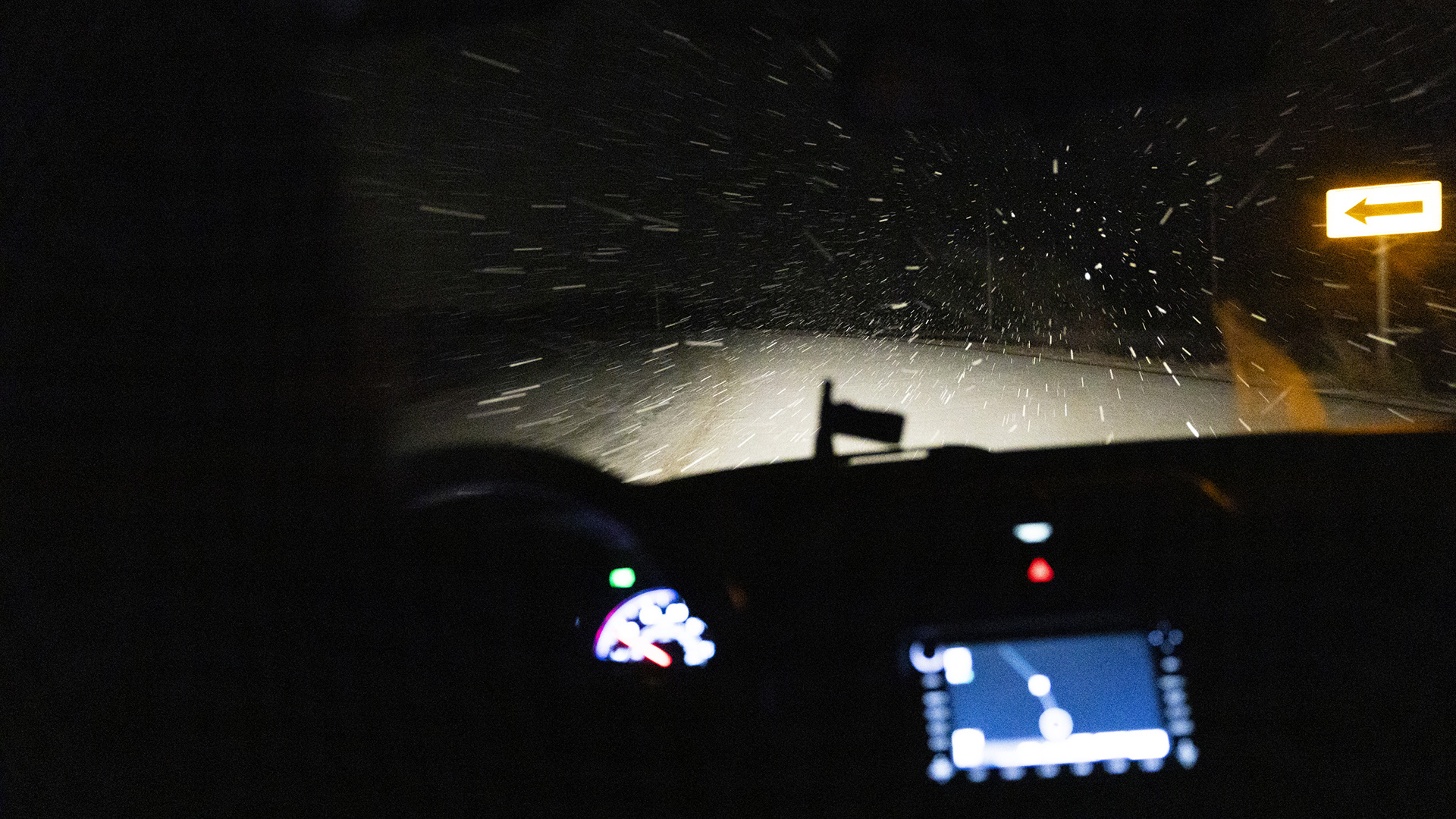 Snow flies toward the windshield of a moving vehicle as its headlights illuminate a traffic sign, as seen inside the cabin with out-of-focus gauges and a dashboard screen showing a map in the foreground.