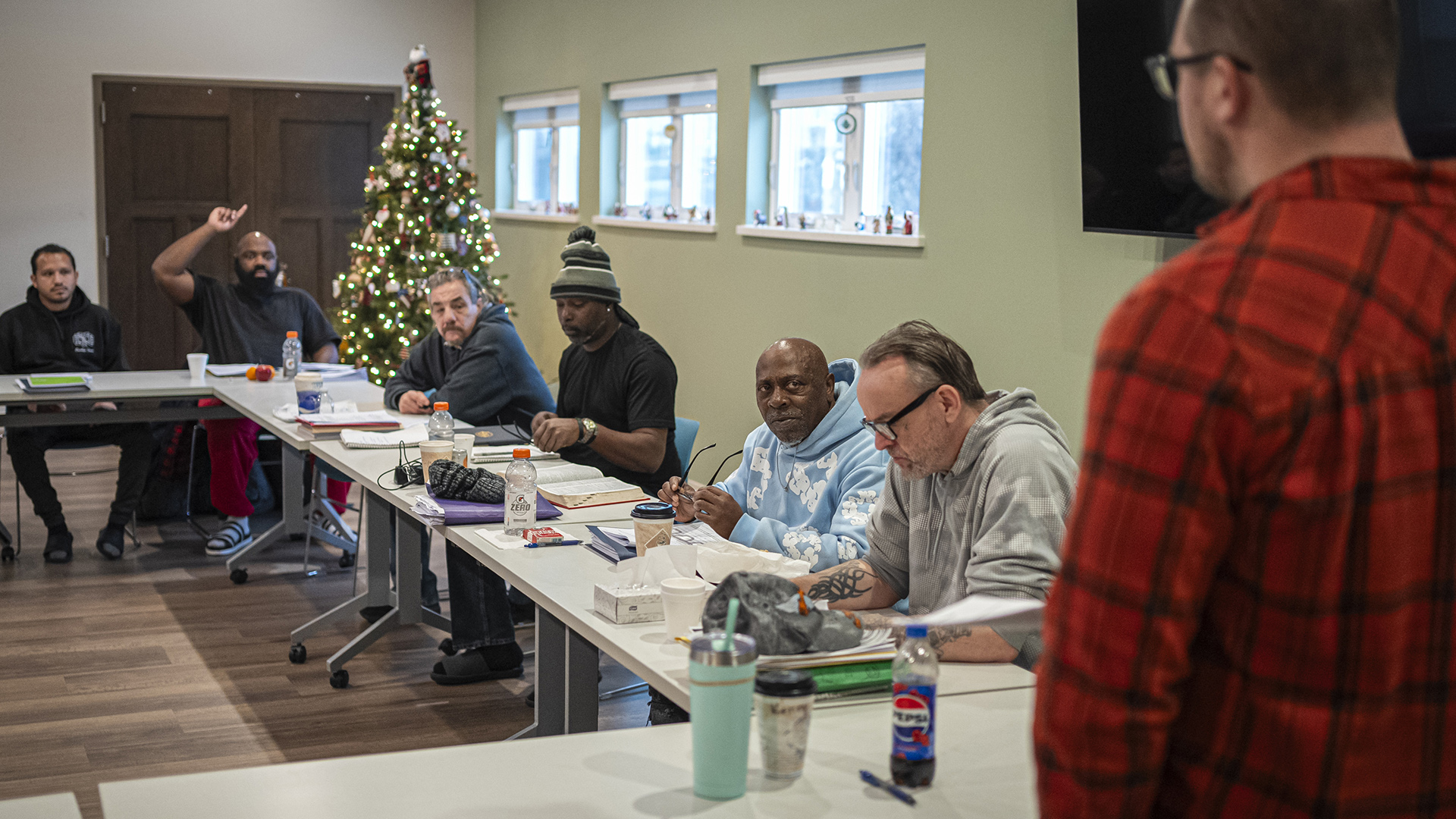 Six men sit on the other edge of a series of rolling tables arranged in a square, with one on the far end raising his hand while facing a man standing in the foreground, in a room with wood double-doors, an illuminated Christmas tree and three windows with raised blinds.