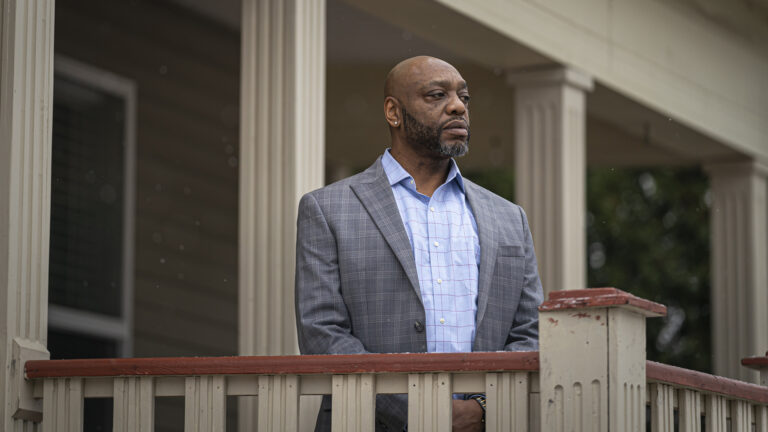 Kenneth Ginlack looks to his left while standing behind a wood railing in front of a building with wood pillars.