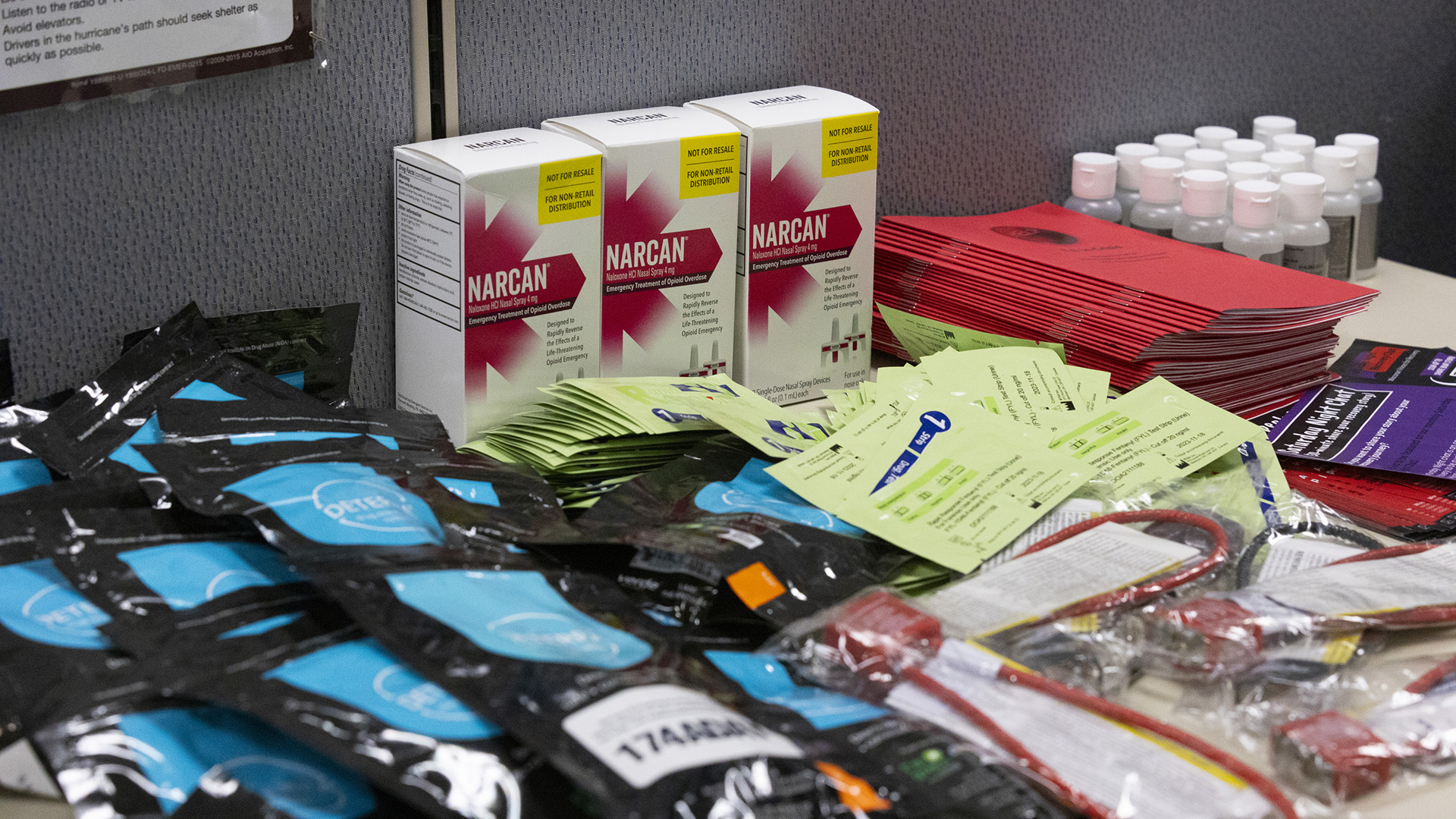 Three cardboard packaging boxes labeled "Narcan," a stack of pamphlets and stacks of other items cover the surface of a table.