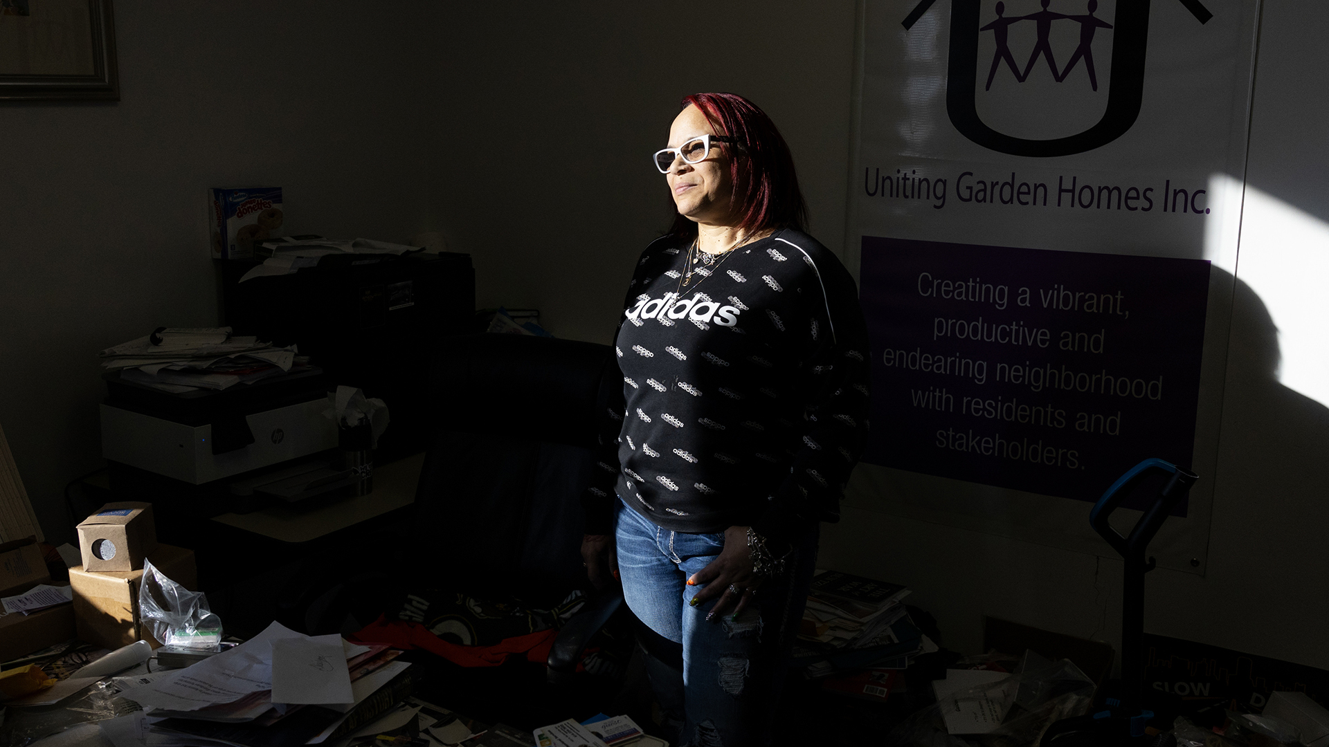 Desilynn Smith is illuminated by sunlight while standing in a shadowed room with stacks of items on the floor and a vinyl sign on a wall reading "Uniting Garden Homes Inc."