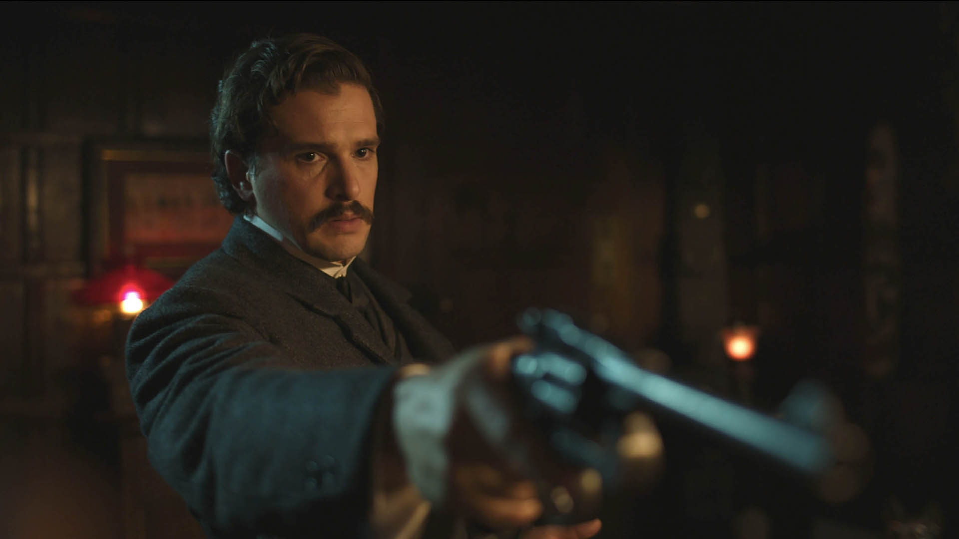 A man with wavy brown hair and a mustache is dressed in a dark Victorian-era suit, pointing a revolver towards the camera with a determined and intense expression.