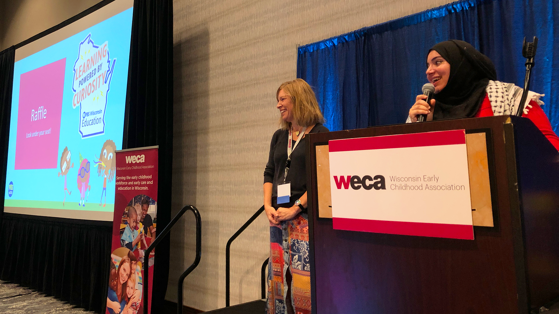A woman in a hijab speaks into a microphone at a podium with a "weca" (Wisconsin Early Childhood Association) sign, while another woman in colorful pants smiles beside her. A large screen in the background displays a "Learning Powered by Curiosity" PBS Wisconsin Education slide, along with a raffle announcement.