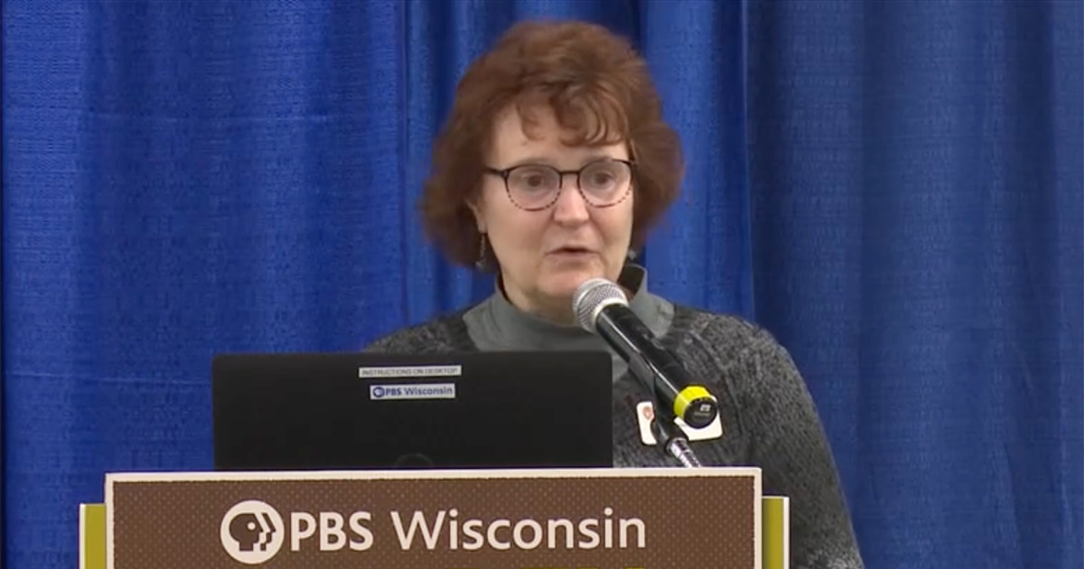 Lisa Johnson speaking into a microphone at a lectern during the 2024 Garden & Landscape Expo, wearing professional clothing in a blue color, with a laptop nearby.