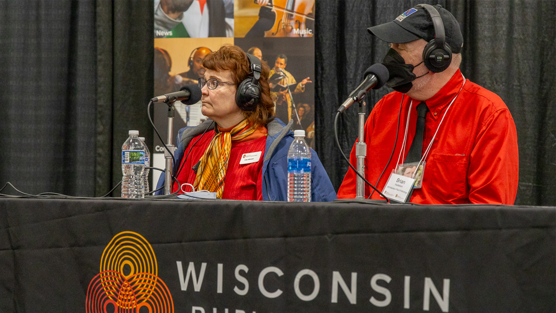 Lisa Johnson (left) and Brian Hudelson (right) sit at a table during WPR's Garden Talk with Larry Meiller live from the 2025 Garden & Green Living Expo with microphones in hand, during an interactive discussion.