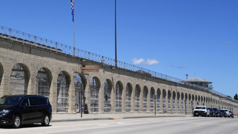 Multiple cars are parked in roadside parking spaces next to a masonry wall with numerous arches enclosed with a metal fence and wood sign reading Waupun Correctional Institution, which stands in front of a tower with a peaked roof in the distance, along with a flagpole with the U.S. and Wisconsin flags and a tall light pole.