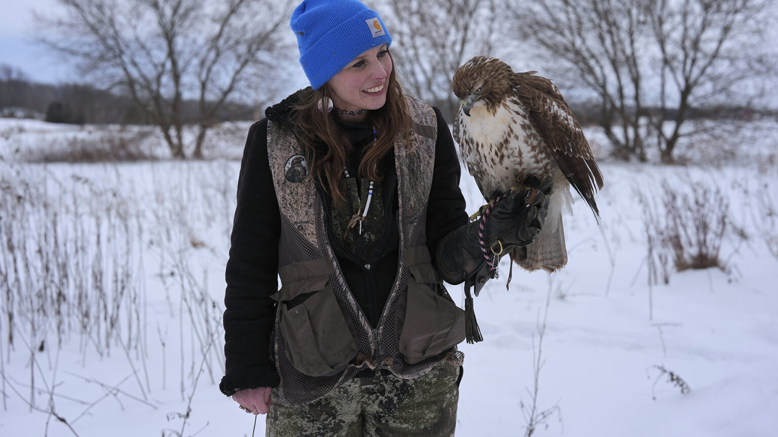 Stephanie Stevens holds a red-tailed hawk in her left hand wearing a falconry gauntlet while standing in a snow-covered field, with leafless trees in the background.