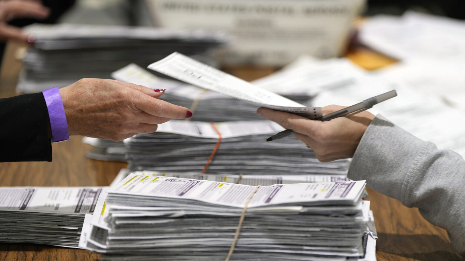 The hand of one election worker that is also holding a pen passes a paper absentee ballot to the hand of another election worker above a table covered with stacks of absentee ballots bound with rubber bands on the surface of a wood table, with a United States Postal Service box in the background.