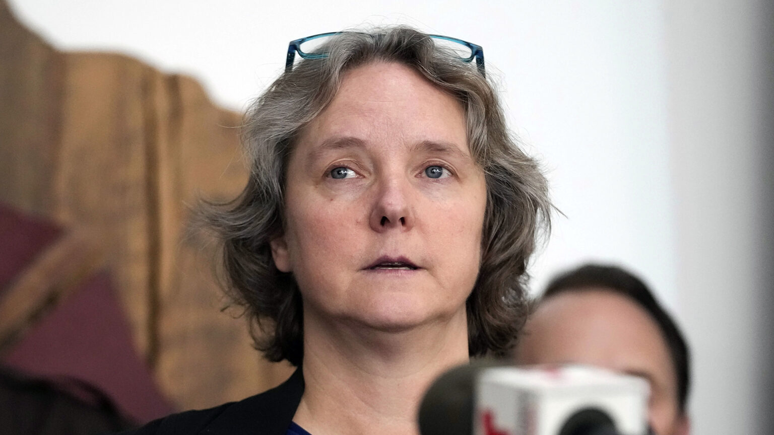 Satya Rhodes-Conway speaks into an out-of-focus microphone with the flag of a media organization, with an out-of-focus person standing behind her.