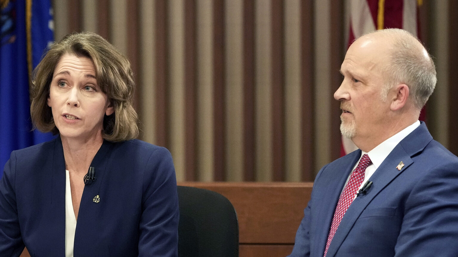 Susan Crawford and Brad Schimel sit next to each other and speak in a room with U.S. and Wisconsin flags next to a wall with vertical wood slats.