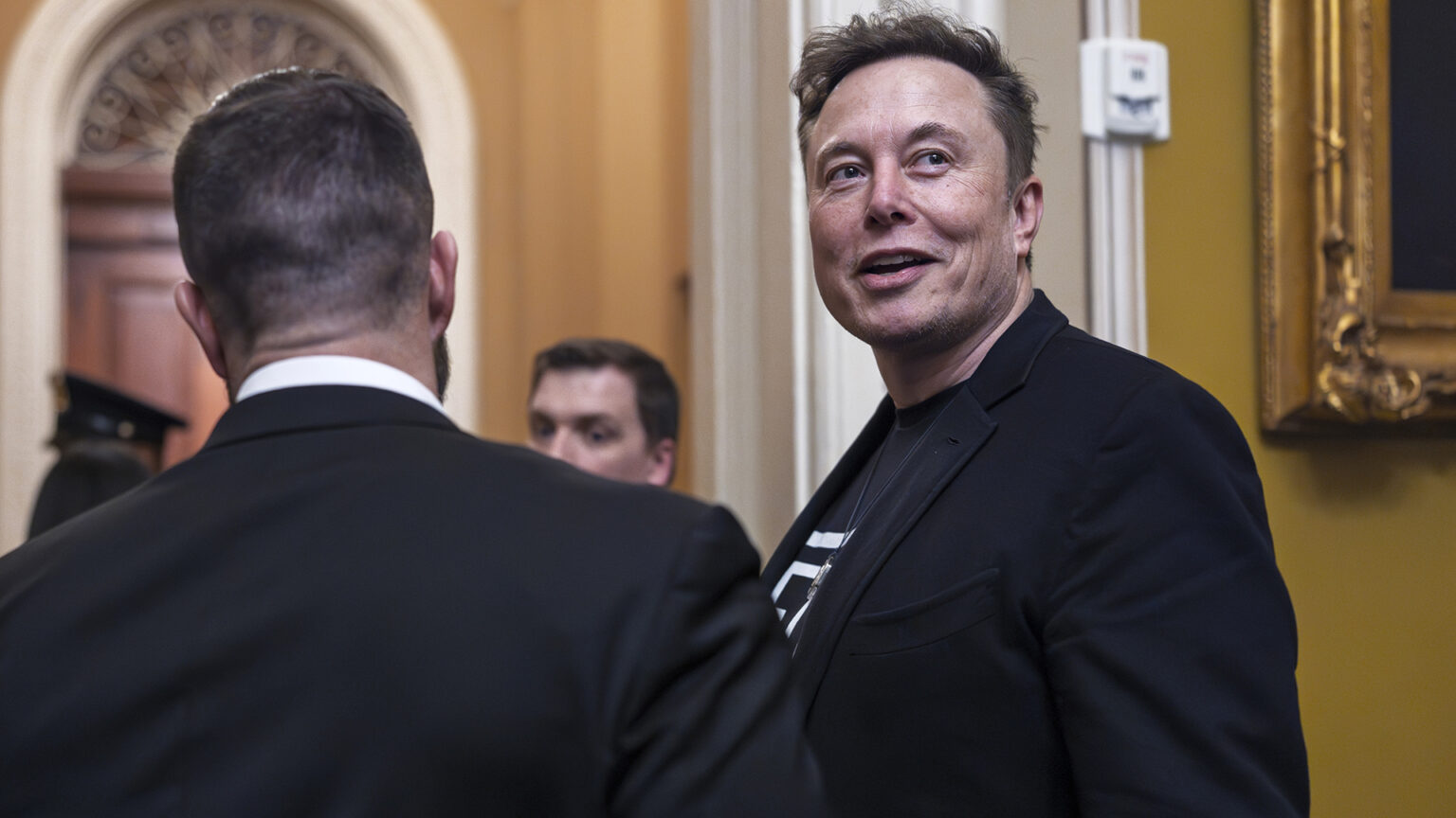 Elon Musk speaks while looking over his left shoulder, with another person standing next to him and others in the background, in a room with arched doorways and a gilt frame of a painting.