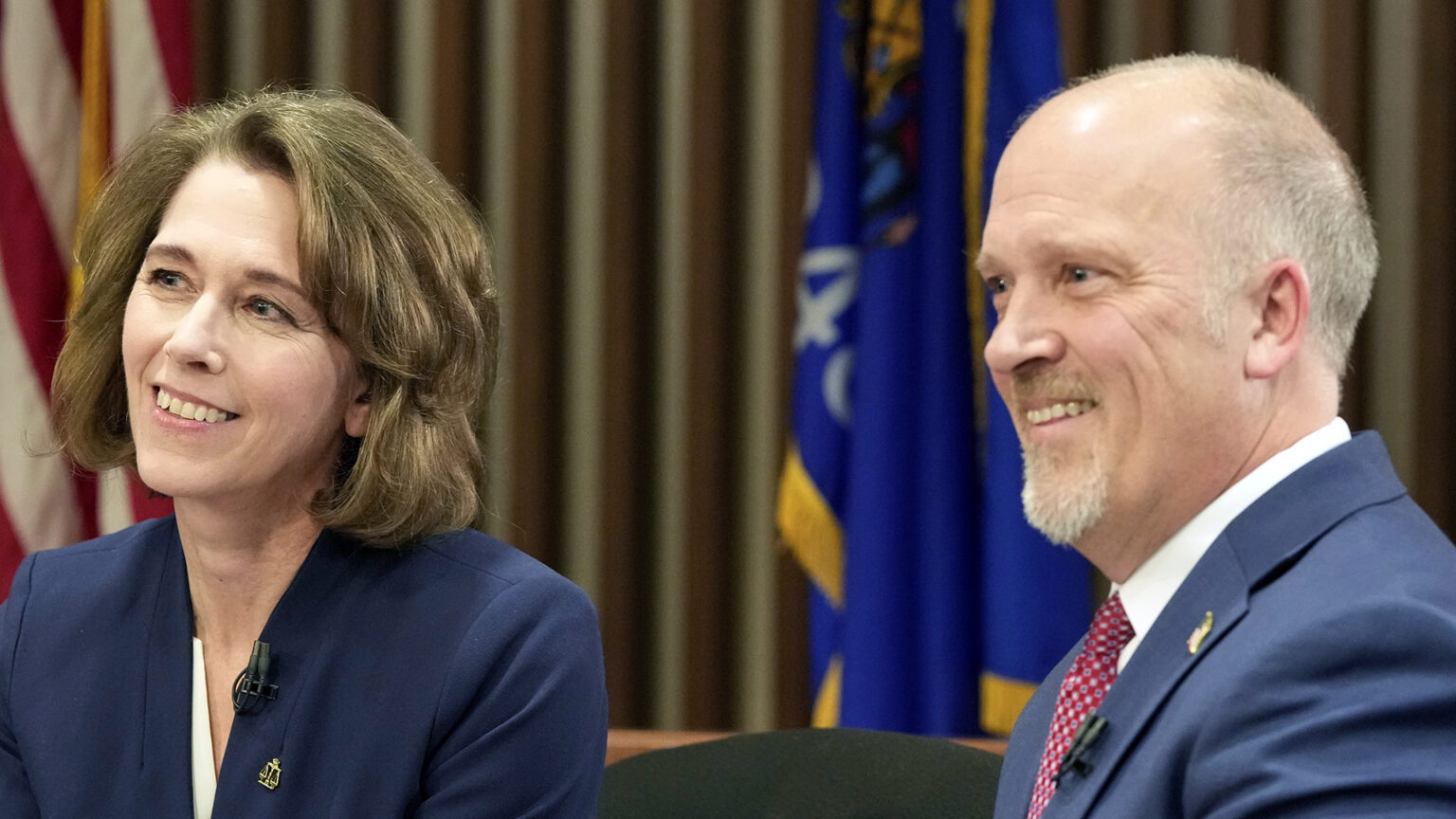 Susan Crawford and Brad Schimel sit next to each other and smile in a room with U.S. and Wisconsin flags next to a wall with vertical wood slats.