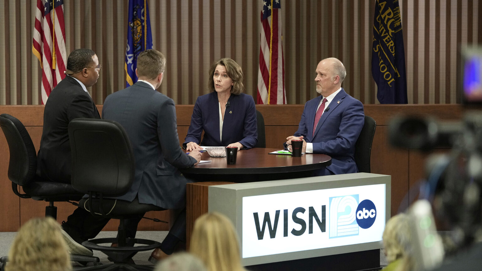 Susan Crawford and Brad Schimel sit next to each other at a round table and face two other people seated on the other side in rolling chairs, with coffee mugs and papers on the table's surface, next to an illuminated sign with the WISN12 wordmark and ABC logo, with out-of-focus seated people and a television camera facing them, in a room with a judicial dais and a row of U.S., Wisconsin and Marquette University Law School flags next to a wall with vertical wood slats.