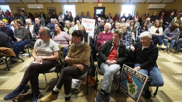 People sit in multiple rows of stackable chairs, with several holding hand-drawn and printed political signs, with a line of people standing along the back wall of a room with low-pile carpet, multiple windows and mini split heat pumps near the ceiling, and a door with a glass window and an illuminated EXIT sign above it.