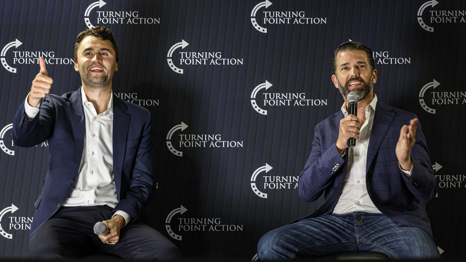 Charlie Kirk makes a thumbs up gesture with his right hand and holds a wireless microphone in his left hand while seated next to Donald Trump Jr., who speaks into a wireless microphone held in his right hand and gestures with his left hand, in front of a backdrop banner with the logo and wordmark of Turning Point Action.