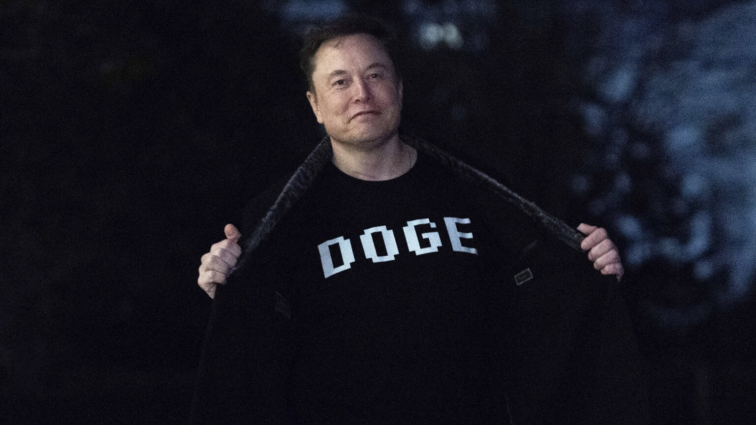 Elon Musk uses both hands to open both sides of a coat to expose a t-shirt with the letters DOGE printed on its front, while standing outdoors in low light at night.