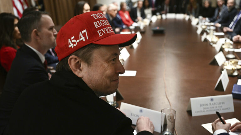 Elon Musk, while wearing a baseball cap with the numerals 45-47 on its right side, looks over his right shoulder while sits at one end of a long, oval table with other people seated around it, with paper name tents, glasses, papers, a cord and other items on its surface.