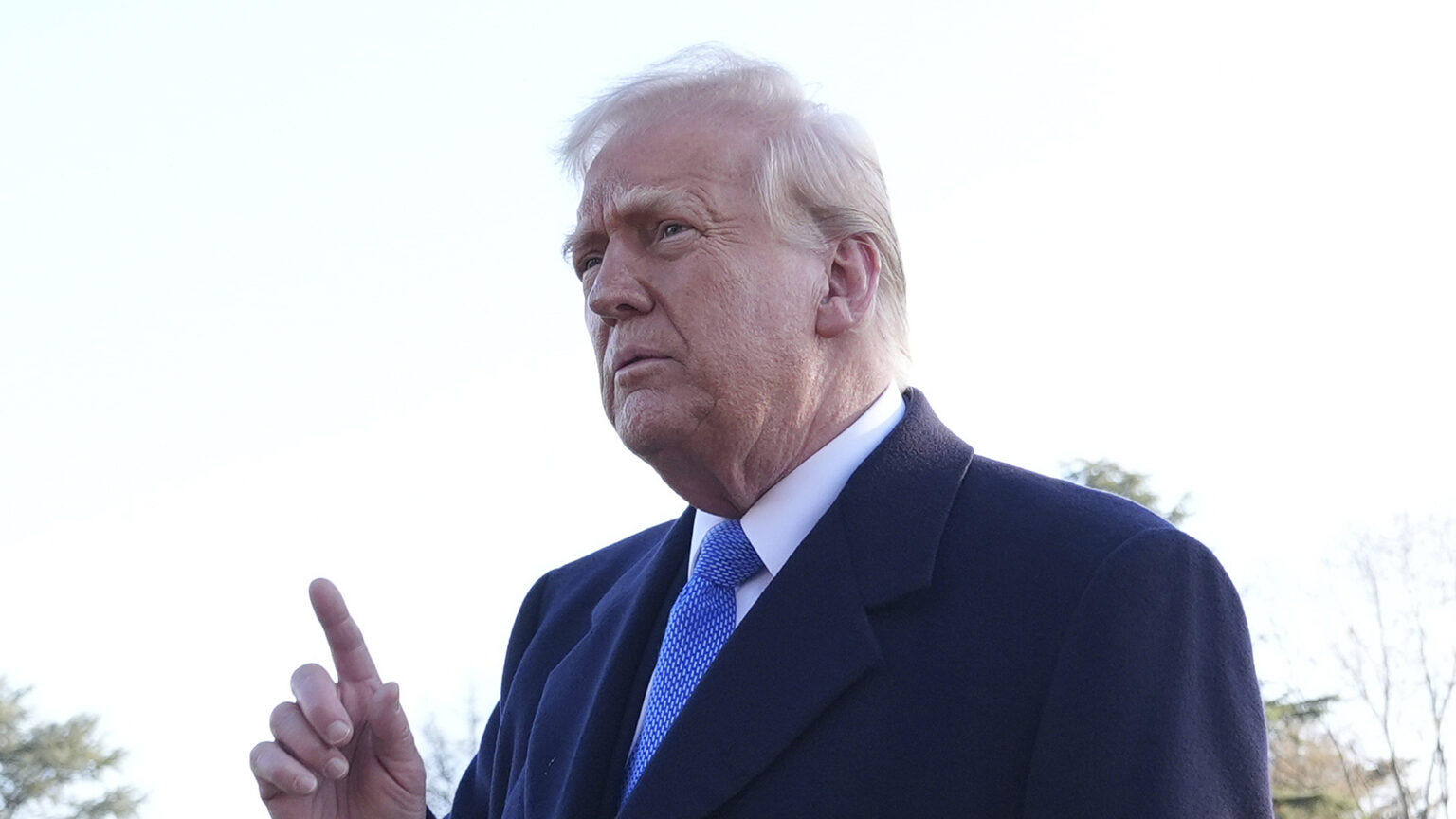 Donald Trump points upwards with the index finger of his right hand while speaking outdoors, with out-of-focus trees in the background.