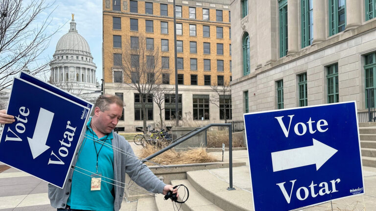 Mike Quieto holds two cardboard yard signs with the words Vote and Votar and an arrow pointing left in his right hand and a pair of headphones in his left hand while standing at the base of a a series of short steps to a multi-story stone masonry building with pillars between its windows on the second floor, with another cardboard sign with the words Vote and Votar and an arrow pointing right in the foreground, with leafless trees, parked bicycles, and a multi-story stone and brick masonry building in the background in front of a building with a dome topped by a statue.