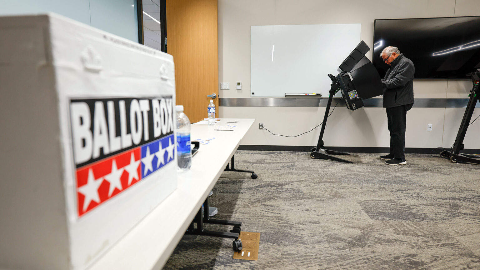 A man stands in front of a mobile metal voting booth with two bases, a stand and multiple folding privacy screens, with another voting both behind him and an open wood door in front of him, in a room with a whiteboard and large monitor on a wall in the background and a low-pile carpet, with an out-of-focus painted box with the words Ballot Box on a table on a side wall.