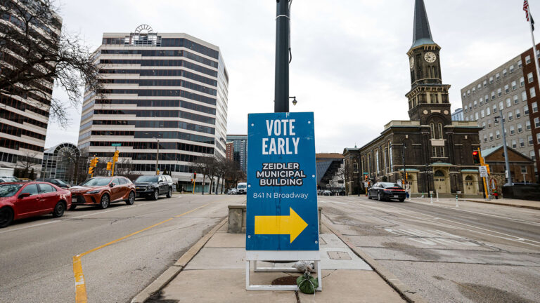 A sandwich-board sign with the words Vote Early and Zeidler Municipal Building, an address and an arrow pointing right stands stabilized with a sandbag on a concrete median between multiple lanes of a street, with driving and parked vehicles on either side, along with a church and multiple multi-story buildings in the background.