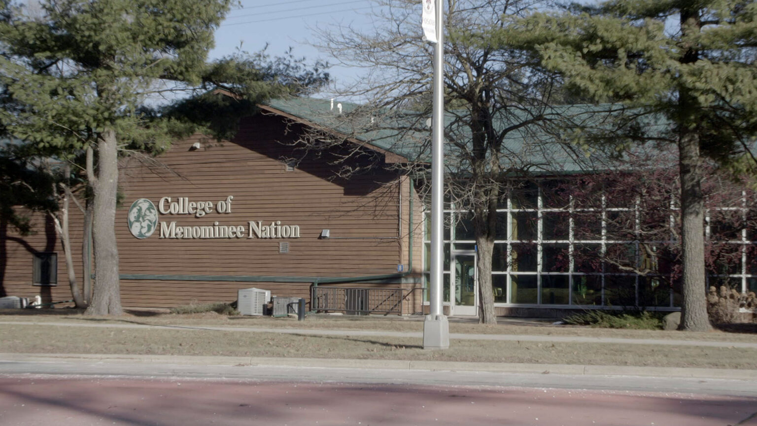 A letter sign reading College of Menominee Nation is attached to the wood-sided wall of a single-story building with a glass-walled atrium and sloped roof, with multiple conifer trees and a metal utility pole standing next to a sidewalk and road in the foreground.