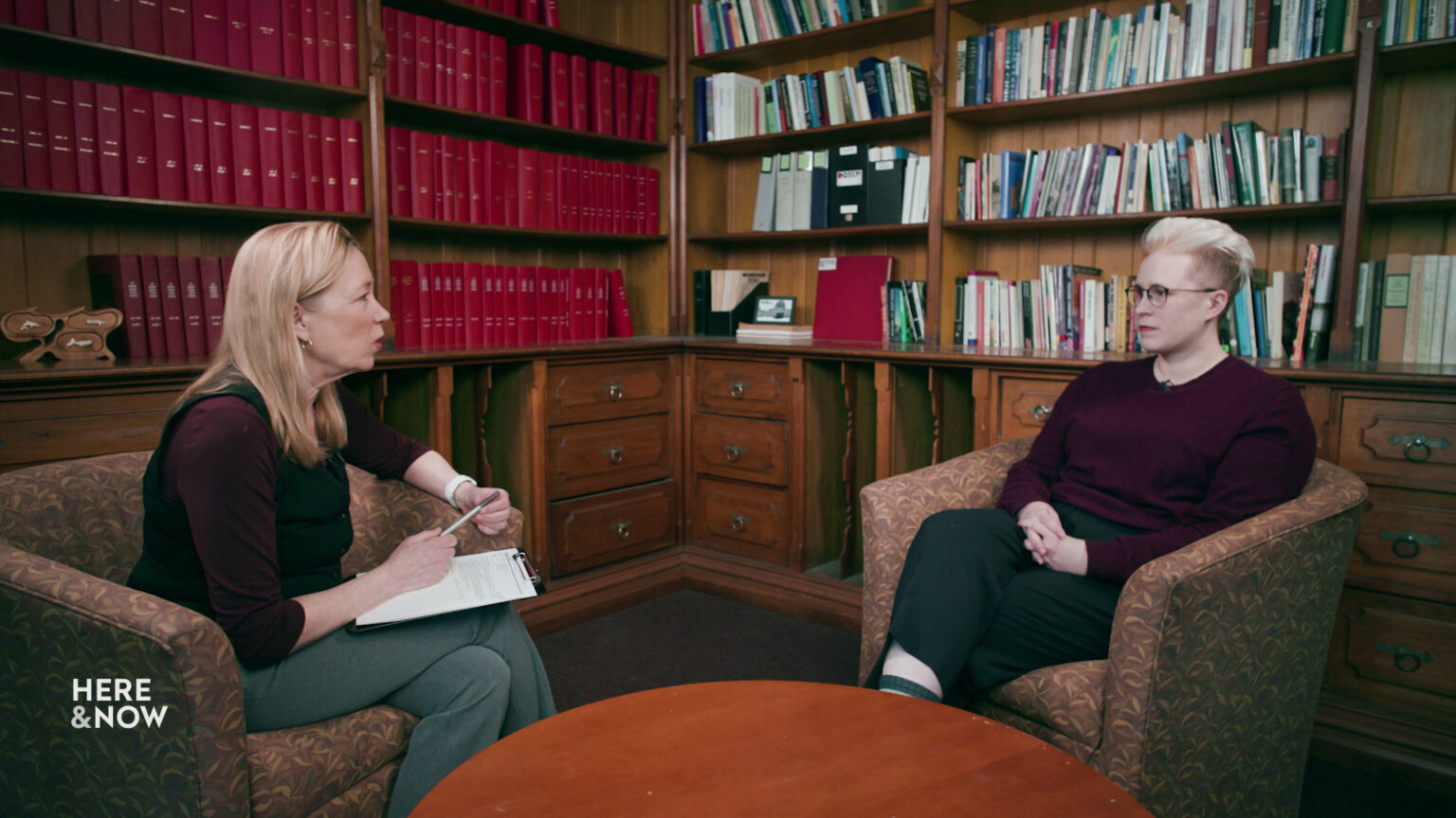 Frederica Freyberg and Mariel Barnes sit in upholstered armchairs facing each other on two sides of a low wood table, in a room with floor to ceiling wood cabinets and bookshelves filled with bound periodicals and books.