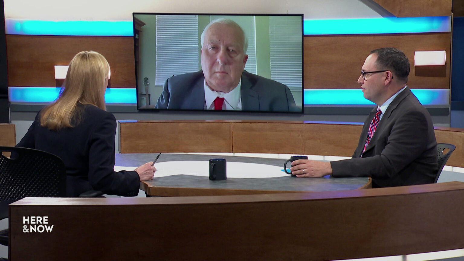 Frederica Freyberg and Jeff Mandell sit on opposite sides of a table on the Here & Now set and face a video monitor showing an image of Rick Esenberg.