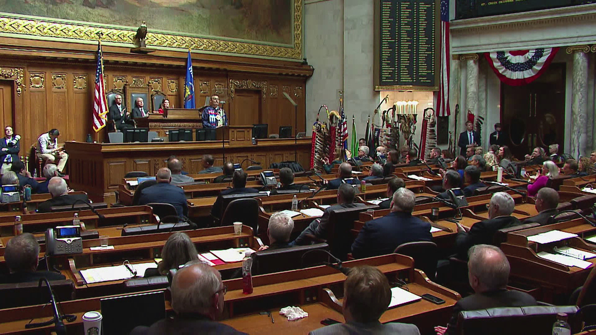 Thomas Fowler stands and speaks into a microphone mounted to a wood podium at the center of a legislative dais while facing two teleprompter mirrors, with three people seated in high-backed wood and leather chairs behind him at a higher level of the dais, faced by more people seated in multiple rows of wood desks, in a room with multiple eagle feather staffs standing to one side, a large painting and taxidermy eagle on the rear wall, an electronic vote register on an adjacent wall, and U.S. flag bunting between marble pillars supporting a second-level seating gallery.