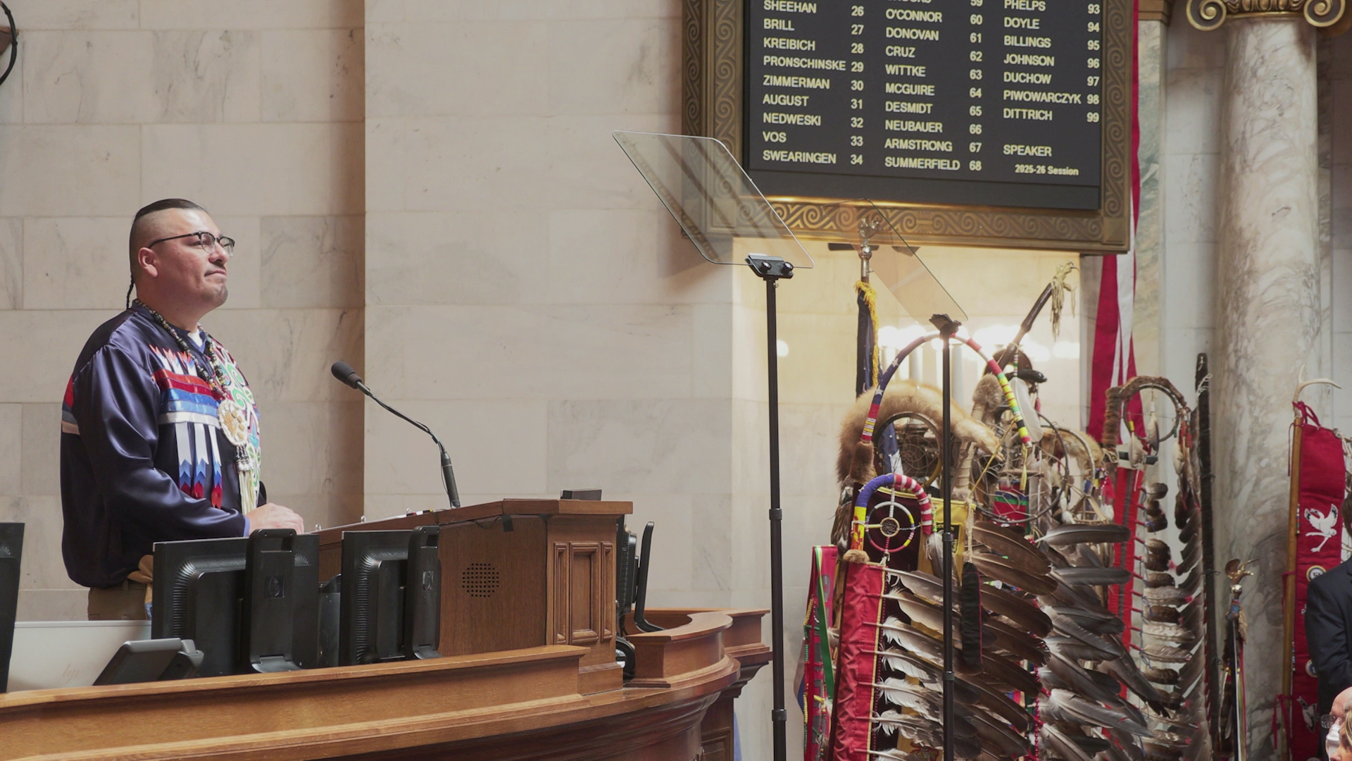 Thomas Fowler stands and speaks into a microphone mounted to a wood podium on legislative dais with multiple computer monitors on either side and while facing two teleprompter mirrors, with multiple eagle feather staffs standing to one side under an electronic vote register, in a room with marble pillars and masonry.