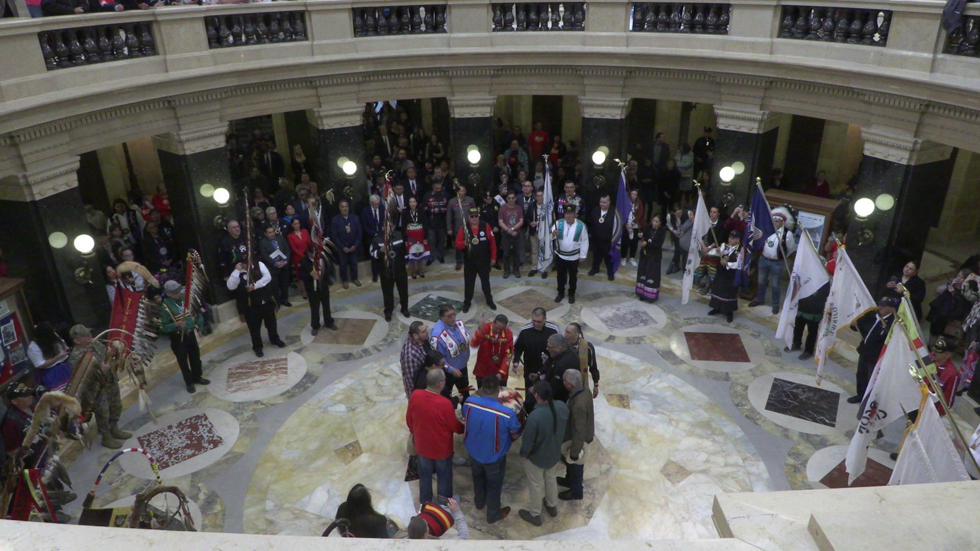 Eleven people stand in a circle around a drum at the center of a rotunda, with a larger concentric circle of people holding flags standing around them and more people standing among square pillars behind them, in a building with a patterns marble floor and a second story balustrade.