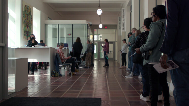 People stand in a line alongside a wall, with its end bending toward a group of tables and chairs where other people are standing and seated, in a high-ceilinged hallway with tile floors, hanging lamps, signs reading Traffic & Parking and Doty St. Exit and a glass wall and doors on the far wall illuminated by an EXIT sign.