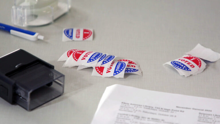 Multiple stickers with the words I Voted Today sit on the surface of a laminate table top next to a self-inking stamp, pen, clear adhesive tape dispenser and a sheet of paper.