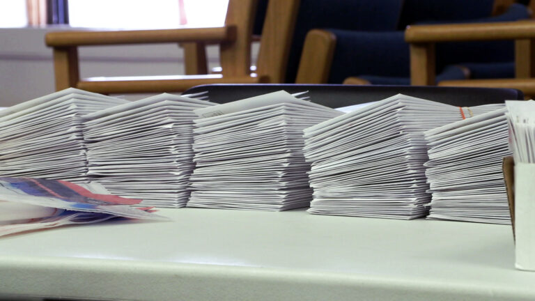 Five stacks of absentee ballot envelopes bound with rubber bands stand in a row on the top of a plastic table surface, with stacks of wood and vinyl-upholstered chairs in the background.