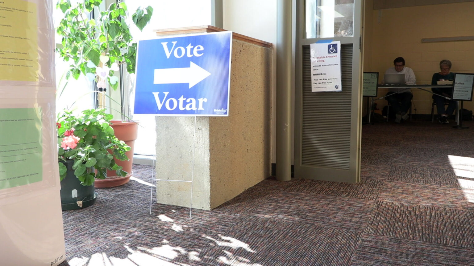 A sign with the words Vote and Votar and an arrow pointing to the right leans up against a concrete half wall next to two potted plants illuminated by sunlight entering through adjacent floor-to-ceiling windows, in a room with a low-pile carpet and two people seated at a folding table in an adjacent space in the background.